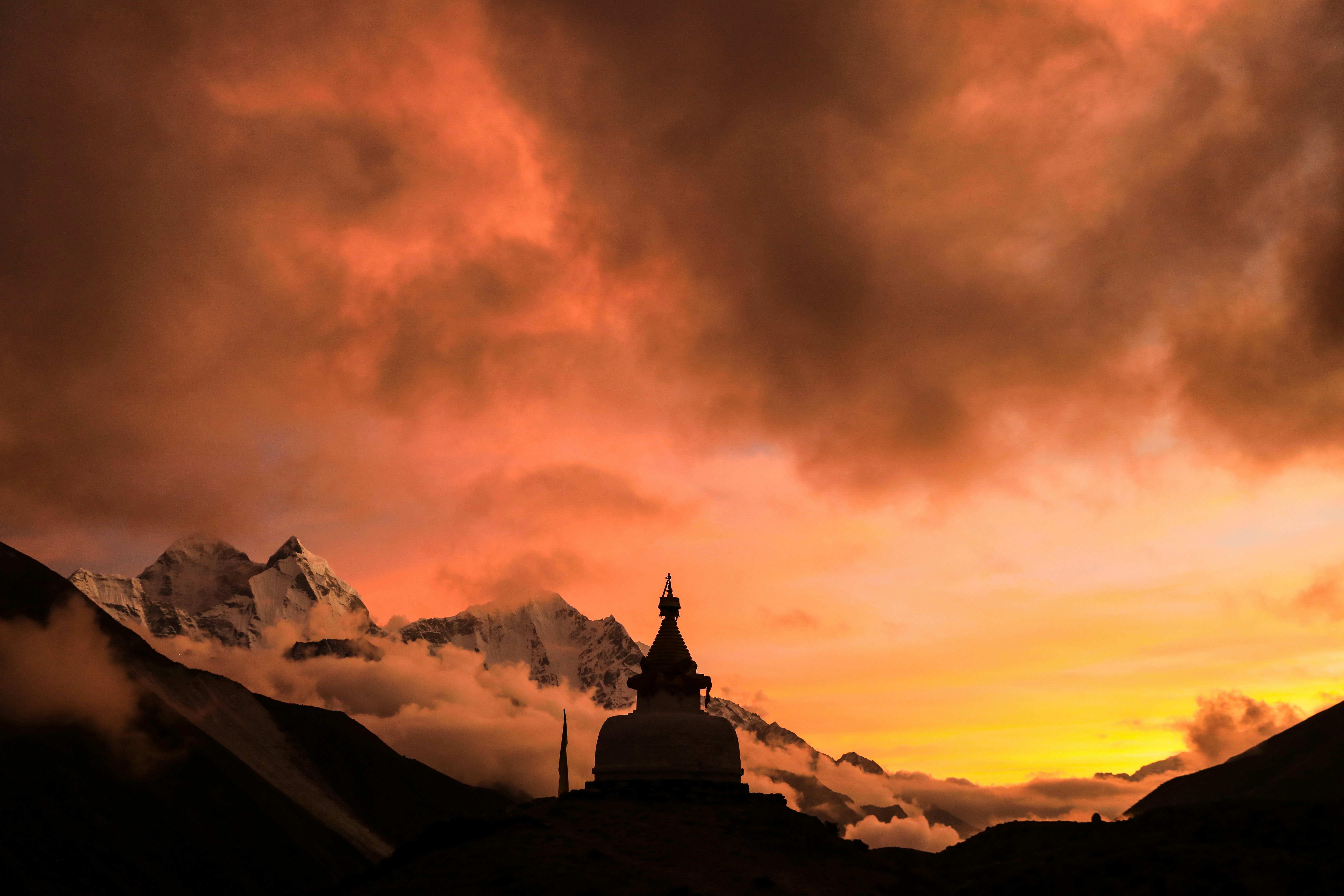 a mountain range with a tower in the foreground and clouds in the background, A dramatic sunset over one of the several stupas dotted throughout the Everest Base Camp trek.
