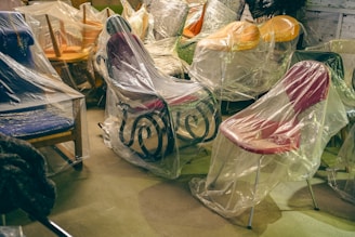 A collection of chairs wrapped in transparent plastic covering, placed in a storage or warehouse-like setting. The variety of chairs includes different styles and colors, such as a blue cushioned chair, a pink plastic chair, and a rocking chair with a black metal frame. The floor is concrete, and the overall atmosphere suggests that the furniture is being protected or stored.