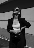 A person is standing confidently against a minimalist architectural background. They are wearing sunglasses, a dark blazer, a white crop top, and high-waisted pants. Their pose exudes confidence and style, with one hand resting on their hip and the other holding a stylish bag.