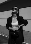 A person is standing confidently against a minimalist architectural background. They are wearing sunglasses, a dark blazer, a white crop top, and high-waisted pants. Their pose exudes confidence and style, with one hand resting on their hip and the other holding a stylish bag.