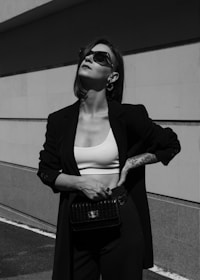 A person is standing confidently against a minimalist architectural background. They are wearing sunglasses, a dark blazer, a white crop top, and high-waisted pants. Their pose exudes confidence and style, with one hand resting on their hip and the other holding a stylish bag.