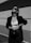 A person is standing confidently against a minimalist architectural background. They are wearing sunglasses, a dark blazer, a white crop top, and high-waisted pants. Their pose exudes confidence and style, with one hand resting on their hip and the other holding a stylish bag.