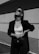 A person is standing confidently against a minimalist architectural background. They are wearing sunglasses, a dark blazer, a white crop top, and high-waisted pants. Their pose exudes confidence and style, with one hand resting on their hip and the other holding a stylish bag.