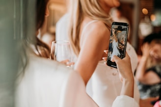 A person is holding a smartphone, capturing a photo of another individual in a light-colored dress. The setting appears to be social with a relaxed atmosphere, as there is a person in the background with a glass of wine.