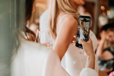 A person is holding a smartphone, capturing a photo of another individual in a light-colored dress. The setting appears to be social with a relaxed atmosphere, as there is a person in the background with a glass of wine.