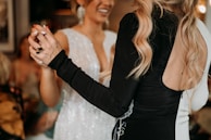 Two women are dancing together. One woman is wearing a white dress with sparkly details, and the other is wearing a black dress with long sleeves and her hair styled in loose waves. The background is blurred, highlighting the focus on the women and their interaction.