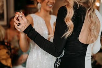 Two women are dancing together. One woman is wearing a white dress with sparkly details, and the other is wearing a black dress with long sleeves and her hair styled in loose waves. The background is blurred, highlighting the focus on the women and their interaction.