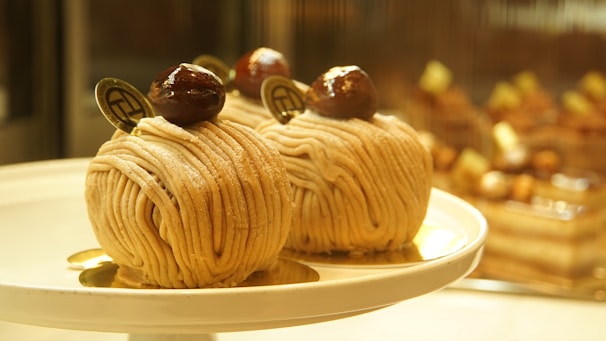 Two intricately crafted desserts with a spiral noodle-like appearance are elegantly presented on a round white plate. Each dessert is topped with a glossy chestnut and a small decorative chocolate piece. The background is softly blurred, showcasing other similar desserts on display.