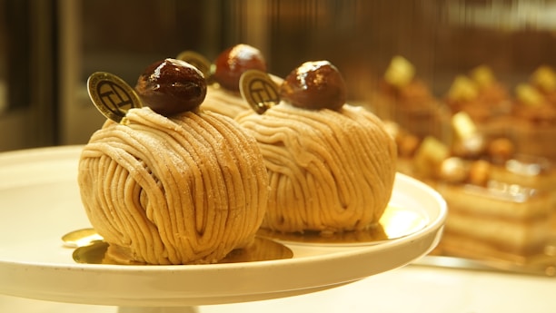 Two intricately crafted desserts with a spiral noodle-like appearance are elegantly presented on a round white plate. Each dessert is topped with a glossy chestnut and a small decorative chocolate piece. The background is softly blurred, showcasing other similar desserts on display.
