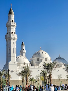 A white mosque with multiple domes and tall minarets under a clear blue sky, surrounded by palm trees. People in colorful clothing walk around in the foreground.