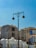 An urban scene featuring modern buildings with Middle Eastern architectural influences. The structures have tall windows and intricate patterns. There are street lamps with ornate designs, and birds are perched on them. The sky is clear with a bright blue hue.