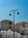 An urban scene featuring modern buildings with Middle Eastern architectural influences. The structures have tall windows and intricate patterns. There are street lamps with ornate designs, and birds are perched on them. The sky is clear with a bright blue hue.