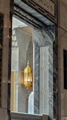 An ornate architectural interior featuring intricate marble patterns and a golden decorative lantern. The design is complemented by Arabic calligraphy and geometric motifs, creating a harmonious blend of colors and textures.