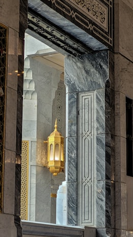An ornate architectural interior featuring intricate marble patterns and a golden decorative lantern. The design is complemented by Arabic calligraphy and geometric motifs, creating a harmonious blend of colors and textures.