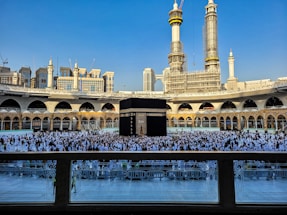 A large crowd of people dressed in white gather around the Kaaba, a black cubic structure with gold embellishments, at the center of a vast open courtyard surrounded by arched buildings. The background features two tall towers under construction along with other ornate architectural elements.