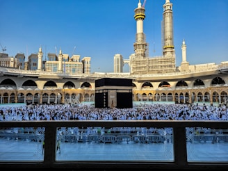 A large crowd of people dressed in white gather around the Kaaba, a black cubic structure with gold embellishments, at the center of a vast open courtyard surrounded by arched buildings. The background features two tall towers under construction along with other ornate architectural elements.