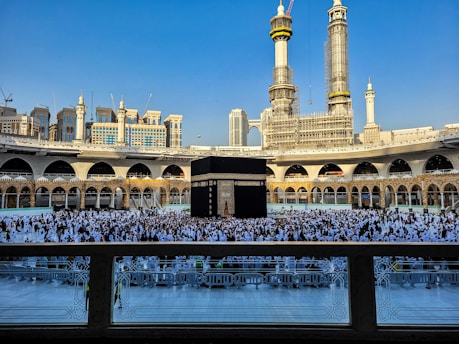 A large crowd of people dressed in white gather around the Kaaba, a black cubic structure with gold embellishments, at the center of a vast open courtyard surrounded by arched buildings. The background features two tall towers under construction along with other ornate architectural elements.