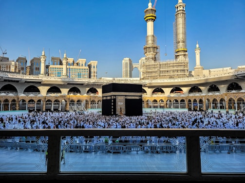 A large crowd of people dressed in white gather around the Kaaba, a black cubic structure with gold embellishments, at the center of a vast open courtyard surrounded by arched buildings. The background features two tall towers under construction along with other ornate architectural elements.