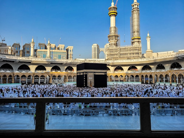 A large crowd of people dressed in white gather around the Kaaba, a black cubic structure with gold embellishments, at the center of a vast open courtyard surrounded by arched buildings. The background features two tall towers under construction along with other ornate architectural elements.