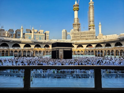 A large crowd of people dressed in white gather around the Kaaba, a black cubic structure with gold embellishments, at the center of a vast open courtyard surrounded by arched buildings. The background features two tall towers under construction along with other ornate architectural elements.