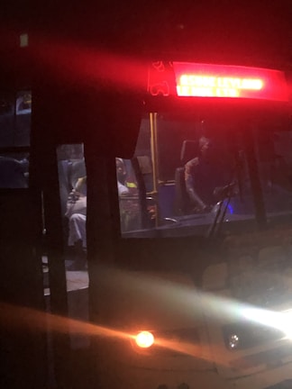A dimly lit bus interior with passengers seated. The bus has a glowing red display showing some text. The headlights and tail lights are on, suggesting it is nighttime.