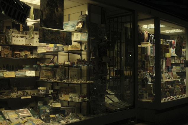 Dimly lit storefront with shelves displaying books, magazines, and various printed materials. Posters, action figures, and other small items are also visible. The glass windows reflect some of the indoor lighting, creating a slightly moody and nostalgic ambiance.