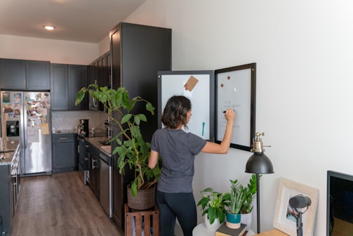 A person is writing on a whiteboard in a modern kitchen. The kitchen has dark cabinets, a refrigerator covered in photos and magnets, and a plant next to the person. There is a microphone and a lamp on a small wooden table nearby.