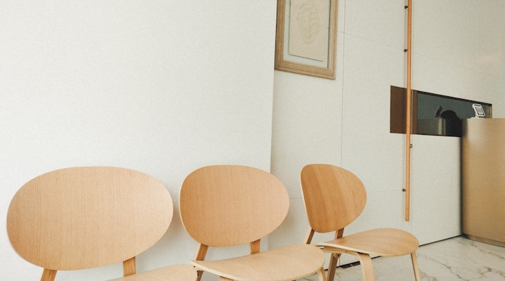 Three light wood chairs with rounded backs are aligned against a white wall. A framed picture mounted on the wall and a modern, minimalistic section with partitions are visible. The floor is likely made of marble. The setting appears organized and calm.