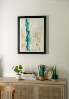 A minimalist interior scene featuring a light wooden cabinet adorned with various decorative items. Above, a framed abstract painting with dominant blue and beige tones hangs on a light-colored wall. On the cabinet, there are small plants in white and dark green pots, a stack of books, and a piece of decorative stone.