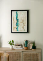 A minimalist interior scene featuring a light wooden cabinet adorned with various decorative items. Above, a framed abstract painting with dominant blue and beige tones hangs on a light-colored wall. On the cabinet, there are small plants in white and dark green pots, a stack of books, and a piece of decorative stone.