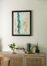 A minimalist interior scene featuring a light wooden cabinet adorned with various decorative items. Above, a framed abstract painting with dominant blue and beige tones hangs on a light-colored wall. On the cabinet, there are small plants in white and dark green pots, a stack of books, and a piece of decorative stone.