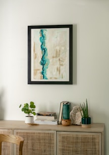 A minimalist interior scene featuring a light wooden cabinet adorned with various decorative items. Above, a framed abstract painting with dominant blue and beige tones hangs on a light-colored wall. On the cabinet, there are small plants in white and dark green pots, a stack of books, and a piece of decorative stone.