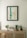 A minimalist interior scene featuring a light wooden cabinet adorned with various decorative items. Above, a framed abstract painting with dominant blue and beige tones hangs on a light-colored wall. On the cabinet, there are small plants in white and dark green pots, a stack of books, and a piece of decorative stone.