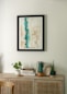 A minimalist interior scene featuring a light wooden cabinet adorned with various decorative items. Above, a framed abstract painting with dominant blue and beige tones hangs on a light-colored wall. On the cabinet, there are small plants in white and dark green pots, a stack of books, and a piece of decorative stone.