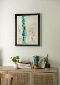 A minimalist interior scene featuring a light wooden cabinet adorned with various decorative items. Above, a framed abstract painting with dominant blue and beige tones hangs on a light-colored wall. On the cabinet, there are small plants in white and dark green pots, a stack of books, and a piece of decorative stone.