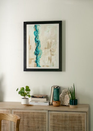 A minimalist interior scene featuring a light wooden cabinet adorned with various decorative items. Above, a framed abstract painting with dominant blue and beige tones hangs on a light-colored wall. On the cabinet, there are small plants in white and dark green pots, a stack of books, and a piece of decorative stone.