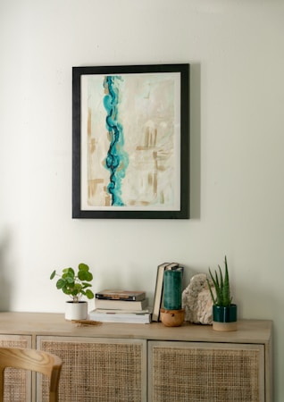 A minimalist interior scene featuring a light wooden cabinet adorned with various decorative items. Above, a framed abstract painting with dominant blue and beige tones hangs on a light-colored wall. On the cabinet, there are small plants in white and dark green pots, a stack of books, and a piece of decorative stone.
