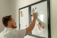A person is writing on a whiteboard with a black marker. The whiteboard has two panels with black frames and is adorned with various notes and images held in place by magnets. The individual has short hair and is wearing a light-colored shirt.