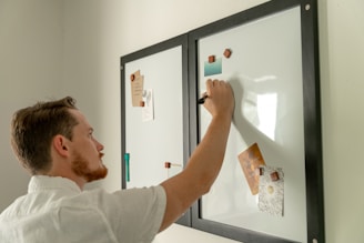 A person is writing on a whiteboard with a black marker. The whiteboard has two panels with black frames and is adorned with various notes and images held in place by magnets. The individual has short hair and is wearing a light-colored shirt.