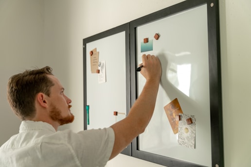 A person is writing on a whiteboard with a black marker. The whiteboard has two panels with black frames and is adorned with various notes and images held in place by magnets. The individual has short hair and is wearing a light-colored shirt.
