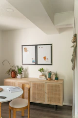 A cozy and bright room corner featuring a light wood cabinet against a cream-colored wall. The cabinet is adorned with potted plants, stacked books, and decorative items. Above it are two small bulletin boards with notes and papers pinned to them. A round table with an open book and chair is positioned in the foreground, and the space is lit with soft natural light.