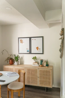 A cozy and bright room corner featuring a light wood cabinet against a cream-colored wall. The cabinet is adorned with potted plants, stacked books, and decorative items. Above it are two small bulletin boards with notes and papers pinned to them. A round table with an open book and chair is positioned in the foreground, and the space is lit with soft natural light.