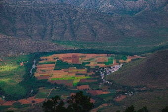 A picturesque valley with a patchwork of agricultural fields in various shades of green and brown, surrounded by hills. A small village with white buildings is nestled at the base of the hills, with a river winding through the landscape.