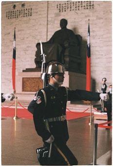 A guard in an elaborate uniform is performing a ceremonial march in front of a large statue seated inside a hall. The guard is wearing a helmet with a reflective surface, and the uniform includes decorative elements such as medals and a belt. Behind the guard, there are national flags displayed next to the statue, which is placed against a wall with inscribed text in a traditional script. The floor is polished, and there are gold stanchions with red ropes creating a barrier around the area.
