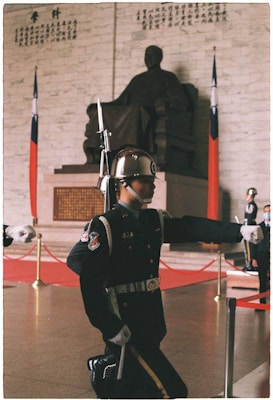 A guard in an elaborate uniform is performing a ceremonial march in front of a large statue seated inside a hall. The guard is wearing a helmet with a reflective surface, and the uniform includes decorative elements such as medals and a belt. Behind the guard, there are national flags displayed next to the statue, which is placed against a wall with inscribed text in a traditional script. The floor is polished, and there are gold stanchions with red ropes creating a barrier around the area.