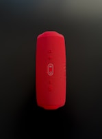 A red cylindrical Bluetooth speaker with a mesh texture, featuring control buttons for power, volume, and wireless connectivity in the center. The speaker is placed vertically against a dark background.