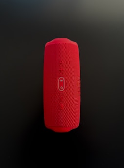 A red cylindrical Bluetooth speaker with a mesh texture, featuring control buttons for power, volume, and wireless connectivity in the center. The speaker is placed vertically against a dark background.