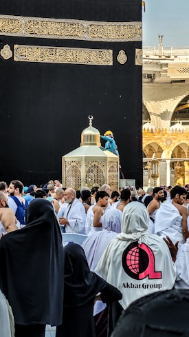 A large group of people gathered in front of a black and gold structure with intricate Arabic calligraphy. The structure, identifiable as part of the Kaaba in Mecca, is surrounded by worshippers dressed in various attires, including white robes. A worker in a blue uniform and yellow helmet is visible on a ladder next to a decorative element.