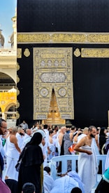 A large gathering of people dressed in white and black garments surrounds the Kaaba, an iconic black cubic structure with intricate gold embroidery, located in a vast open area. The crowd appears to be engaged in a religious ceremony, with many people facing towards the Kaaba in reverence. The structure's prominent door is visible, adorned with elaborate golden patterns and Arabic calligraphy.