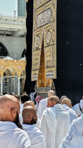 A group of people dressed in white robes gather around the Kaaba, a cube-shaped structure draped in black cloth with gold Arabic script. The scene captures the spiritual atmosphere of a holy place with architectural elements visible in the background.